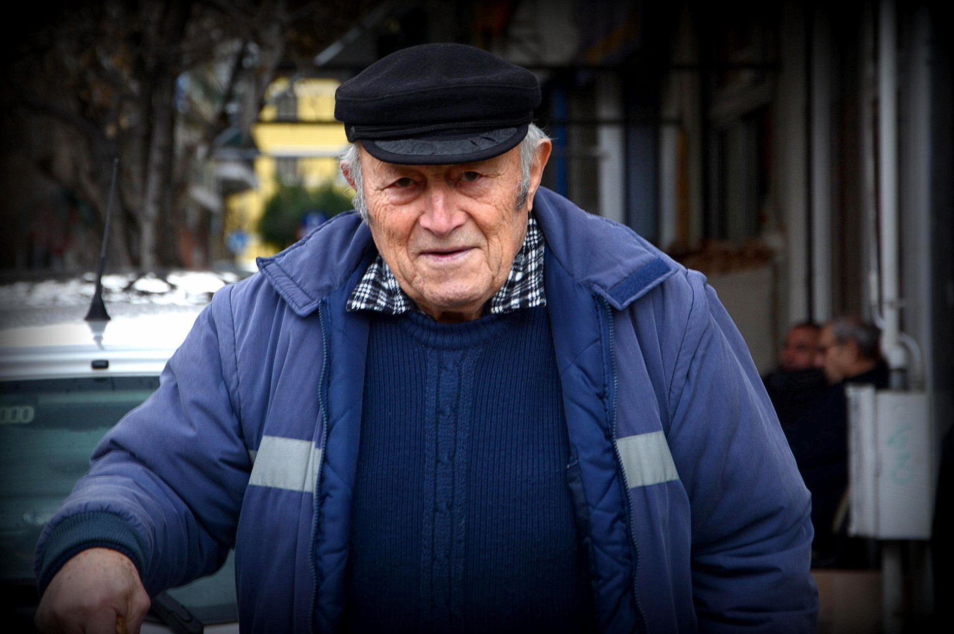 Close-up portrait of an elderly man wearing a blue jacket and black cap, captured outdoors.