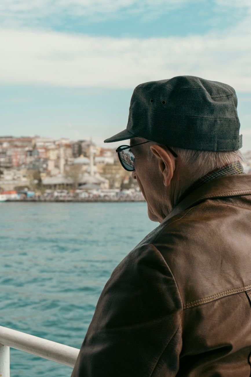 A senior man in a cap and jacket looks over the water towards the cityscape from a boat.
