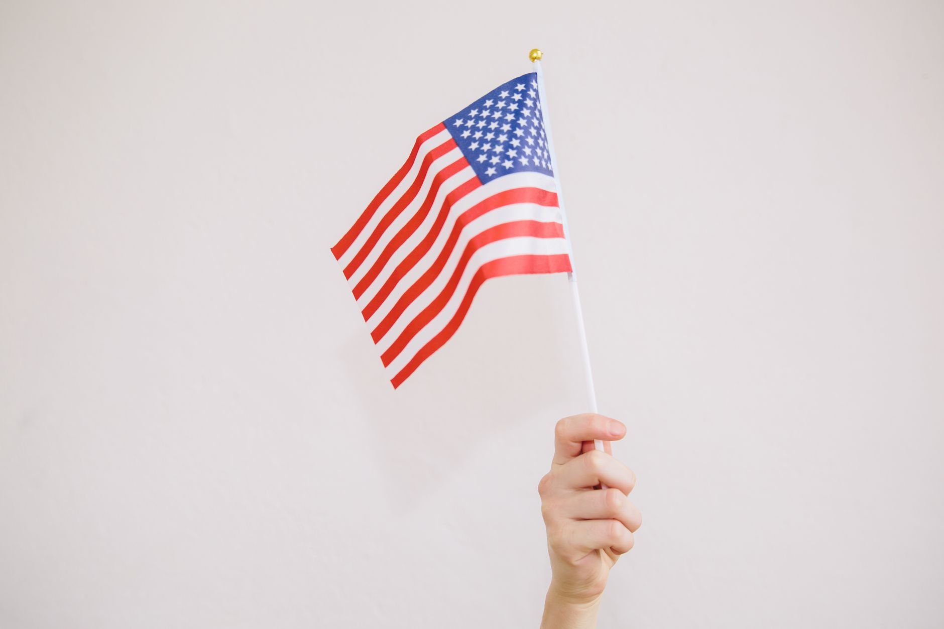 Close-up of a hand holding a waving American flag on a neutral background.